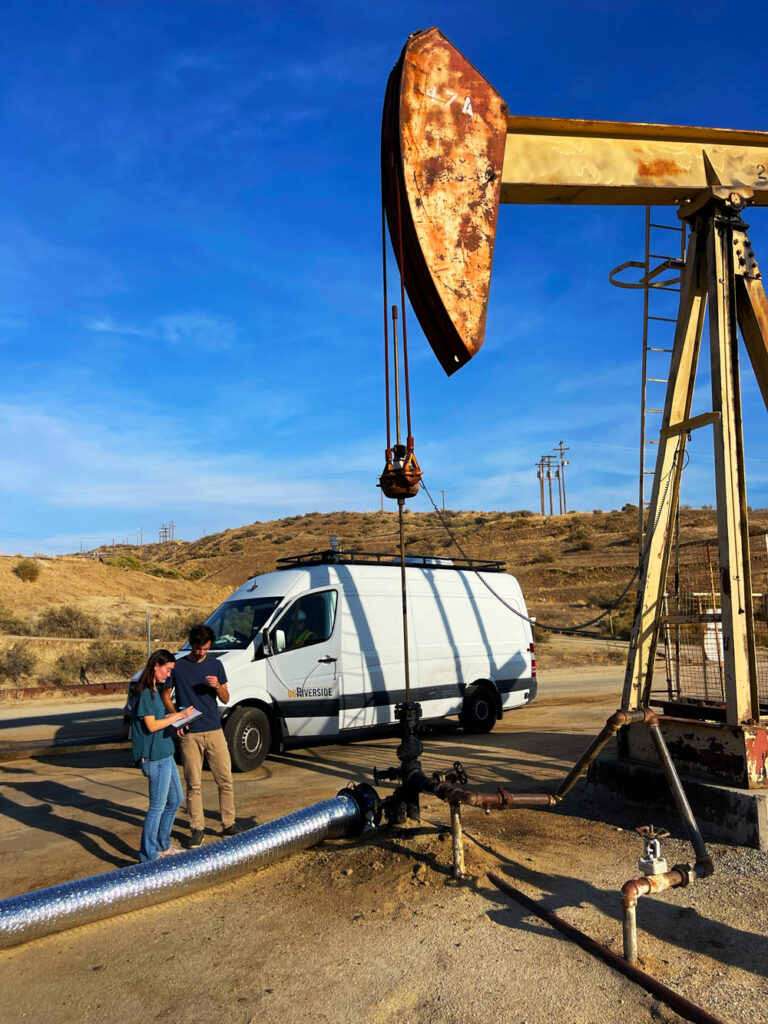 Image of two researchers next to an oil well, looking at a clipboard, standing in front of a foil pipe and a white van labelled UC Riverside